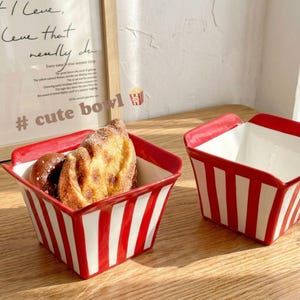May include: Two ceramic bowls with a red and white striped design. One bowl contains baked goods. The bowls have a rectangular shape with a red rim. The background includes a framed print and the text "# cute bowl".