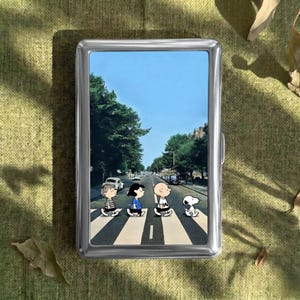 Pode incluir: Um porta-cigarros de metal prateado com uma imagem colorida dos personagens de Peanuts atravessando uma rua, que lembra a capa do álbum Abbey Road dos Beatles. A imagem mostra Charlie Brown, Lucy, Linus e Snoopy andando em uma faixa de pedestres.