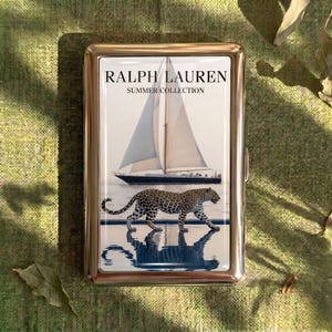 Puede incluir: Un estuche de cigarrillos rectangular de metal dorado. El estuche presenta una imagen impresa de un velero en el agua y un leopardo. El texto "RALPH LAUREN SUMMER COLLECTION" está impreso en la parte superior.