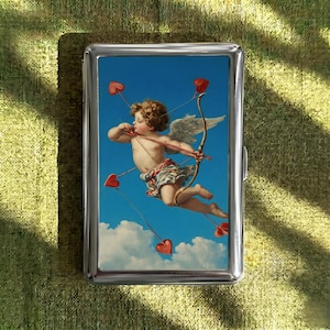 Op de afbeelding: Een zilveren metalen sigarettenetui met een schilderij van Cupido die pijlen in de vorm van harten afschiet. Het kunstwerk toont een cherubijnfiguur met vleugels tegen een blauwe lucht met wolken. De etui heeft een glanzende, metalen afwerking.