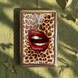 May include: A gold-toned metal cigarette case featuring a leopard print design. The case has a glossy image of red lips with gold and diamond tooth gems. The case is rectangular and has a hinged opening.