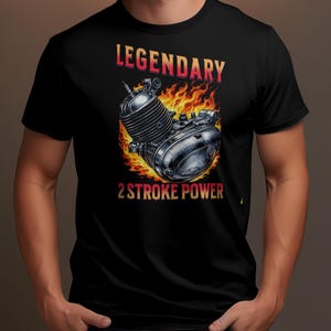 Könnte beinhalten: Schwarzes T-Shirt mit einer detaillierten Illustration eines Motorradmotors in Flammen. Der Text "LEGENDARY" steht über dem Motor und "2 STROKE POWER" darunter. Das Design verwendet Rot-, Orange- und Silbertöne.