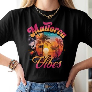 Könnte beinhalten: Ein schwarzes T-Shirt mit einem Grafikdruck eines Sonnenuntergangs über einem Strand mit Palmen und dem Text Mallorca Vibes in einer rosa-orangen Farbverlaufsschrift. Das Design umfasst einen kreisförmigen Rahmen und kleine Sterne.