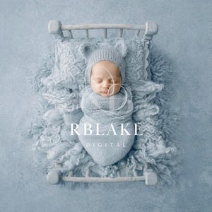 May include: A newborn baby swaddled in a light blue wrap and hat with bear ears, lying on a miniature bed. The bed is set on a fluffy, light blue blanket. The image has a soft, dreamy aesthetic.