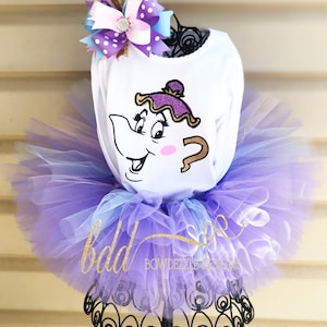 Puede incluir: Una camiseta blanca de manga larga con un diseño de tetera de dibujos animados, combinada con un tutú morado y azul en capas. Un lazo decorativo con lunares morados y detalles dorados está encima. El texto "bdd BOWDEZINE DESIGNS" es visible.