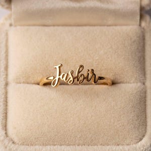 Può includere: Un anello color oro con il nome Jasbir scritto in carattere corsivo. L'anello è centrato all'interno di un portagioie foderato in velluto beige, caratterizzato da una finitura metallica lucida e un design a fascia sottile.