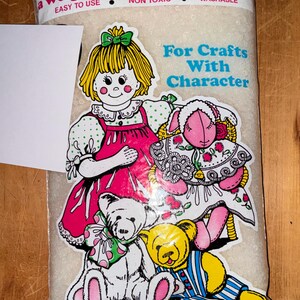 May include: A clear plastic bag filled with white poly-pellets, a weighted stuffing material. The label features a cartoon illustration of a child with yellow hair, a pink dress, and two teddy bears. Text includes "poly-pellets" and "For Crafts With Character."