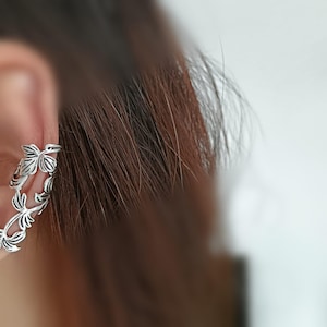 May include: Silver ear cuff with a detailed leaf design. The cuff wraps around the ear, displaying intricate leaf and vine patterns. The jewellery is designed to be worn on the ear without requiring a piercing.