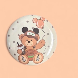 May include: A circular pinback button featuring a light brown teddy bear wearing a brown cap with ears, brown overalls, and holding a boba tea drink. The bear holds a peach-colored balloon, and the background is cream with small gray stars.