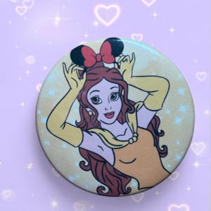 May include: A circular pinback button featuring a colorful illustration of a brown-haired character wearing a yellow dress and long yellow gloves, holding a pair of black mouse ears with a red bow above their head against a light yellow background.