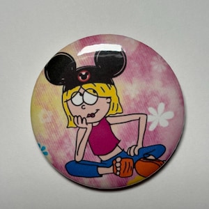 May include: A circular pinback button featuring a cartoon illustration of a blonde character wearing black mouse ears with a red center emblem. The character wears a pink crop top and blue pants, set against a pink and yellow tie-dye background.