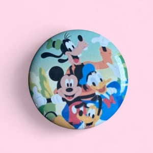 May include: A circular pinback button featuring a colorful graphic illustration of Mickey Mouse, Donald Duck, Goofy, and Pluto. The characters are depicted together in a cheerful pose against a light blue background with green foliage elements.