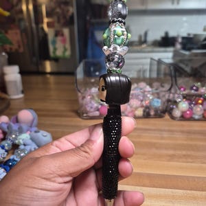 以下が含まれることがあります： 黒いテクスチャのバレルと、黒、銀、緑の積み重ねられたビーズで構成されたトッパーを備えた装飾的なペン。トッパーには、ペンのバレルの上に配置された、横顔の黒髪のキャラクターの小さなフィギュアが含まれています。