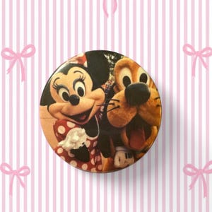 May include: A circular pinback button featuring a colorful graphic of Minnie Mouse and Pluto. Minnie Mouse is wearing her signature red and white polka dot dress, while Pluto is shown with a wide, happy expression. The design is vibrant and clear.