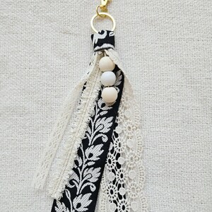 May include: A handmade keychain with a gold-coloured clasp and ring. The tassel features black and white patterned fabric, cream-coloured lace, and three wooden beads. The design incorporates floral and lace patterns.