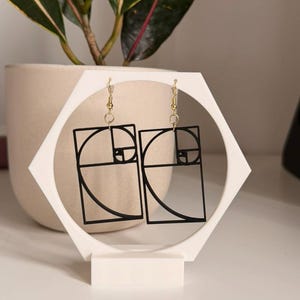 Può includere: Orecchini geometrici neri con ganci color oro, esposti su un supporto esagonale bianco. Gli orecchini presentano un design a sequenza di Fibonacci. Il supporto è su una superficie bianca, con una pianta in vaso sullo sfondo.