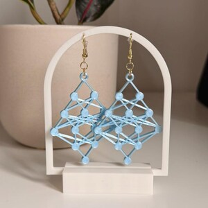 May include: A pair of light blue geometric earrings with gold-coloured ear wires. The earrings are displayed on a white stand. The earrings have a network design. The stand is in front of a potted plant.