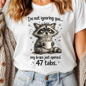 以下が含まれることがあります： コーヒーカップを持った漫画のアライグマが描かれた白いTシャツ。「I'm not ignoring you... my brain just opened. 47 tabs.」と書かれています。アライグマは疲れた表情をしています。