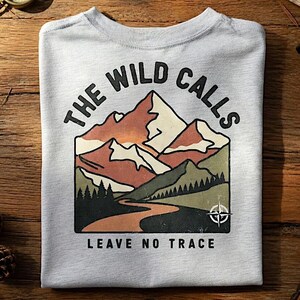 Op de afbeelding: Een lichtgrijs t-shirt met een afbeelding van bergen, een rivier en de tekst "THE WILD CALLS LEAVE NO TRACE". Het ontwerp gebruikt bruin, groen en wit. Het shirt is op een houten oppervlak gevouwen.