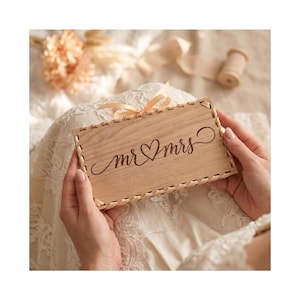 Könnte beinhalten: Eine hellfarbene Holzplakette mit den Worten "mr & mrs" in eleganter Schrift, mit einem Herzsymbol zwischen den Wörtern. Die Plakette hat einen gewebten Rand und wird von einer Person gehalten.