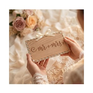 Könnte beinhalten: Ein helles Holzschild mit den Worten "mr + mrs" in Schreibschrift. Das Schild hat oben ein cremefarbenes Band und einen dekorativen, genähten Rand. Das Schild wird vor einem weichen, verschwommenen Hintergrund mit Blumen gehalten.