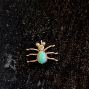 Puede incluir: Un colgante de araña plateada con una piedra ovalada de color turquesa en el centro. La araña tiene seis patas y una cabeza detallada. El colgante es probablemente una joya.