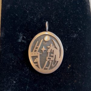 Puede incluir: Colgante de plata con forma ovalada, con un diseño detallado de una figura estilizada, una escalera, una estrella y una luna. El colgante tiene un lazo en la parte superior para una cadena y está sobre un fondo oscuro.