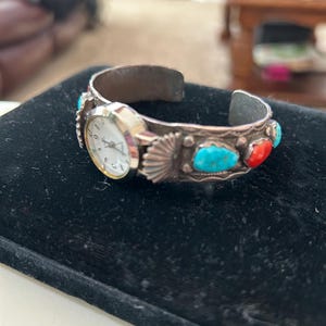 Puede incluir: Reloj de pulsera plateado con esfera blanca y detalles de piedras turquesas y rojas. El reloj presenta un diseño decorativo de concha y está colocado sobre una superficie negra. Una joya única.