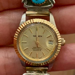 Puede incluir: Un reloj vintage con esfera y correa doradas, con una banda decorativa plateada y dorada con detalles turquesa y rojos. La esfera del reloj muestra la marca "GRIEN". El reloj es resistente al agua.