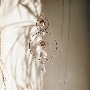 May include: A hanging suncatcher featuring two thin gold-toned metal rings, a central diamond-shaped gold charm with a crescent moon cutout, and multiple clear faceted glass crystals suspended vertically on a delicate gold-toned metal chain.