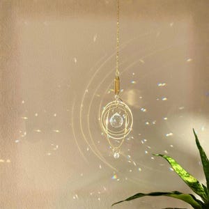 Pode incluir: Um apanhador de sol de metal dourado com uma conta de cristal transparente facetada suspensa dentro de anéis circulares concêntricos. O ornamento pende de uma delicada corrente dourada, projetando reflexos de luz na área circundante.