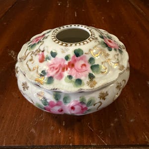 May include: A small, round, antique porcelain trinket box with a lid. The box is white with pink roses, green leaves, and gold accents. The lid has a hole in the center. The box is sitting on a wooden surface.