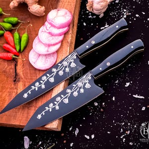 May include: Two black kitchen knives with silver floral engravings on the blades and black handles with metallic rivets. The knives feature the text ECB Elite Cut Blades. The blades are sharp and pointed, designed for culinary tasks in a kitchen.