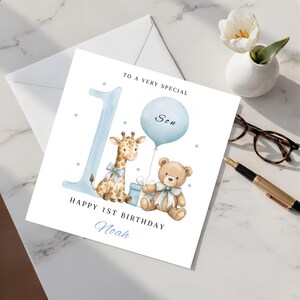 May include: A square birthday greeting card featuring a large light blue number one, a watercolor giraffe, a teddy bear, a blue balloon, and a gift box. The text reads TO A VERY SPECIAL Son, HAPPY 1ST BIRTHDAY, and the name Noah in blue script.