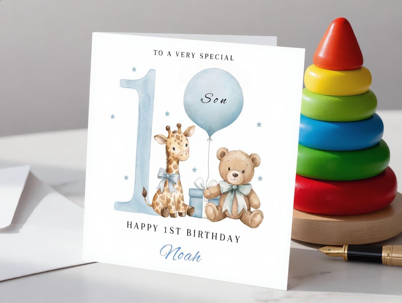 May include: A square white greeting card featuring a large blue number one, a watercolor giraffe with a blue bow, a light blue balloon labeled Son, a brown teddy bear with a blue bow, and a gift box. Text reads To a very special, Happy 1st Birthday, and Noah.