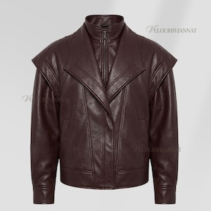 Puede incluir: Chaqueta de cuero burdeos oscuro con cuello alto y cremallera completa. La chaqueta presenta un diseño único con hombros estructurados y un corte entallado. La marca "Velourbyjannat" es visible.