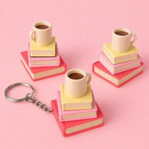 Könnte beinhalten: Ein Schlüsselanhänger mit einer Miniatur-Kaffeetasse auf einem Stapel bunter Bücher. Die Bücher sind in Rosa, Gelb und Rot gehalten. Die Kaffeetasse ist beige, und der Schlüsselanhänger hat einen silbernen Ring und eine Kette. Der Hintergrund ist einfarbig rosa.