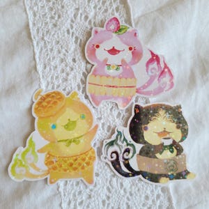May include: Three die-cut stickers featuring stylized cat-like creatures with holographic accents. One is pink with a strawberry motif, one is yellow with an acorn hat, and one is black with a coffee cup, all wearing patterned waist wraps.