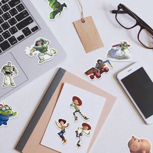 May include: A collection of colorful stickers featuring characters from the animated film Toy Story. The stickers include Buzz Lightyear, Woody, Jessie, and other characters. The stickers are arranged on a white surface with a laptop, phone, and glasses.
