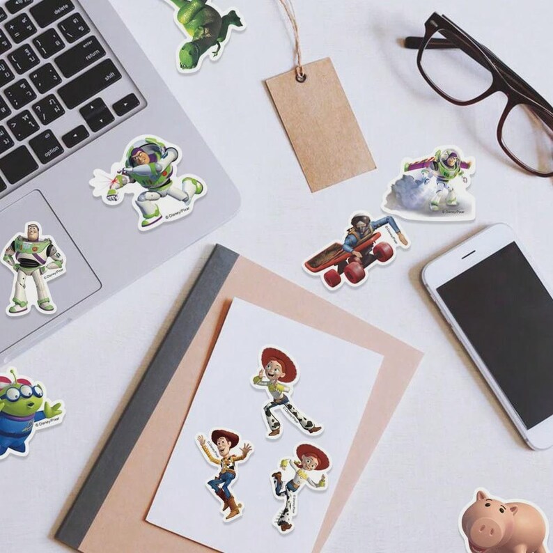 May include: A collection of colorful stickers featuring characters from the animated film Toy Story. The stickers include Buzz Lightyear, Woody, Jessie, and other characters. The stickers are arranged on a white surface with a laptop, phone, and glasses.