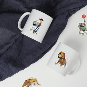 May include: White ceramic mugs featuring colorful stickers of characters from the animated film Toy Story. The stickers include Woody, Buzz Lightyear, and Slinky Dog. The mugs are set against a dark blue fabric background.