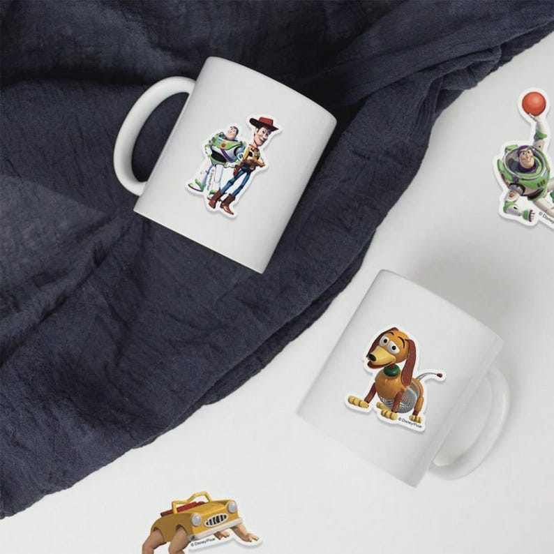 May include: White ceramic mugs featuring colorful stickers of characters from the animated film Toy Story. The stickers include Woody, Buzz Lightyear, and Slinky Dog. The mugs are set against a dark blue fabric background.