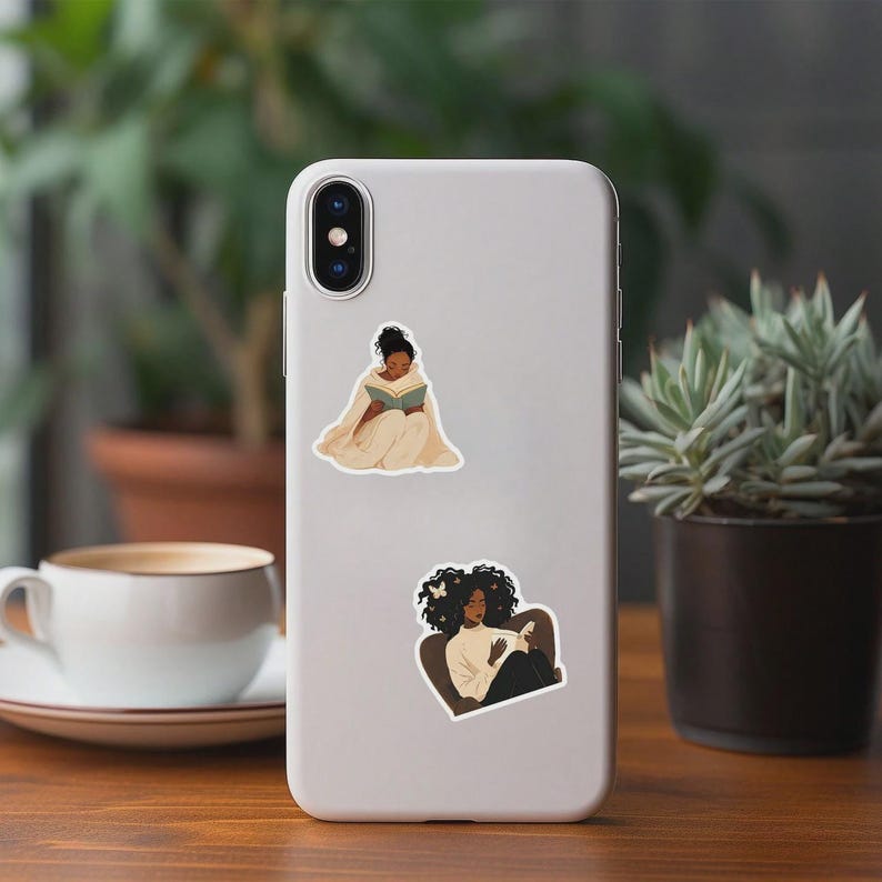 May include: A light gray phone case with two stickers. One sticker shows a person reading a book while wrapped in a blanket. The other sticker shows a person reading a book while sitting in a chair.