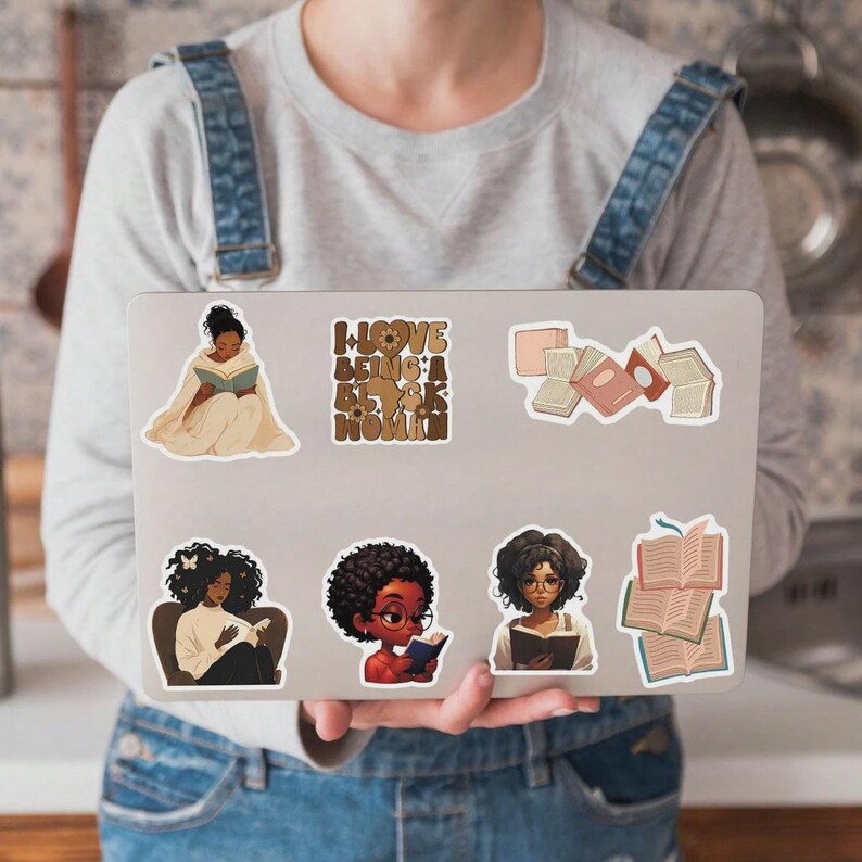 May include: A laptop covered in stickers featuring illustrations of Black women reading books. The stickers have a warm color palette and include the text "I Love Being a Black Woman."