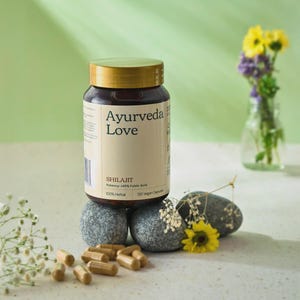 Puede incluir: Un frasco marrón con tapa dorada con la etiqueta "Ayurveda Love" que contiene cápsulas de Shilajit. La etiqueta indica 100% herbal y 120 cápsulas veganas. El frasco descansa sobre piedras grises, con cápsulas esparcidas debajo y un pequeño jarrón de flores.