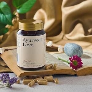 Puede incluir: Un frasco marrón con tapa dorada con la etiqueta "Ayurveda Love" se encuentra sobre un libro abierto. El frasco contiene 120 cápsulas veganas. Varias cápsulas están esparcidas alrededor del libro, junto con una flor rosa y una pequeña piedra gris.