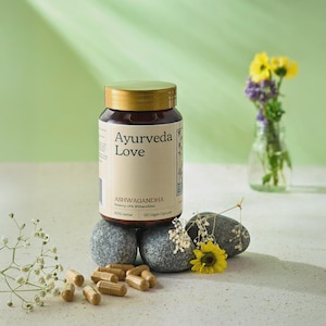 Puede incluir: Una botella de vidrio marrón con tapa dorada, etiquetada "Ayurveda Love" y "Ashwagandha", se encuentra sobre piedras grises. La botella contiene 120 cápsulas veganas. También se ven flores amarillas y moradas en un jarrón y cápsulas.