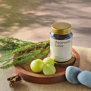 Puede incluir: Una botella de vidrio oscuro con tapa dorada con la etiqueta "Ayurveda Love" se encuentra en una bandeja de madera con fruta verde y follaje. La botella contiene 120 cápsulas veganas. Dos piedras grises lisas están a la derecha.