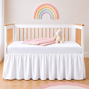 Könnte beinhalten: Ein weißes Kinderbett aus hellem Holz mit weißer Matratze, einer einfarbigen rosa Decke und einem beigen Teddybären. Das Bett hat vertikale Stäbe und einen weißen Bettrüschen-Volant. Über dem Bett befindet sich eine pastellfarbene Regenbogen-Wanddeko.