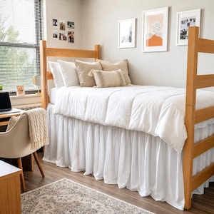 May include: A wooden loft bed frame with a white comforter and a white bed skirt. The bed is styled with two large white pillows, two beige square pillows, and one rectangular beige accent pillow. The bed is positioned in a room with light walls.
